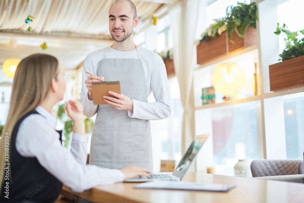 Fototapeta premium Smiling waiter writing down order of client in notepad during working day