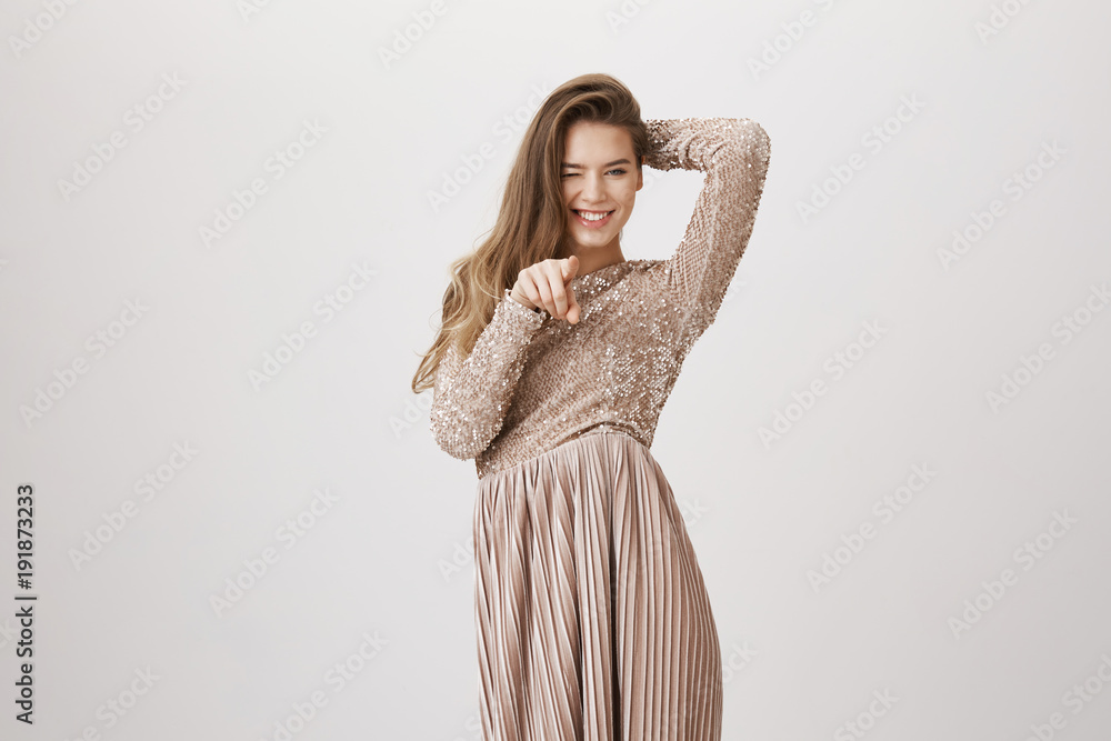 Portrait of charming european adult businesswoman in fashionable evening dress, pointing at camera and winking, flirting and standing over gray background. Bridesmaid having fun on wedding