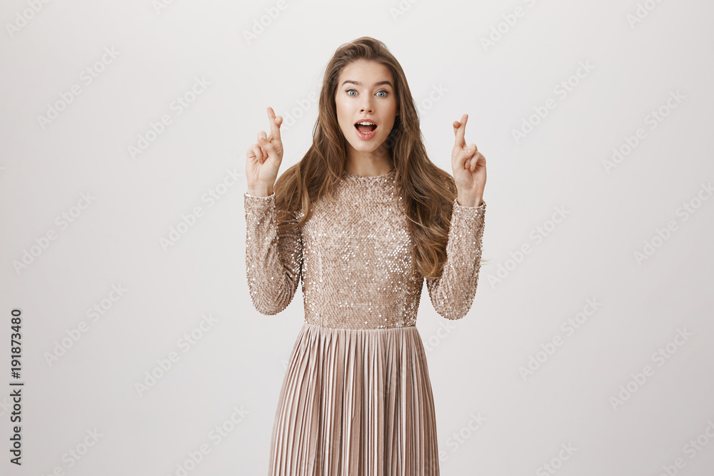 Portrait of gorgeous european model in dress raising hands with crossed fingers and glancing at camera with opened mouth, wishing, hoping that something will come true, standing over gray background Portrait of gorgeous european model in dress raising hands with crossed fingers and glancing at camera with opened mouth, wishing, hoping that something will come true, standing over gray background
