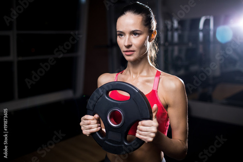 Strong young woman holding disc from barbell and exercising in gym