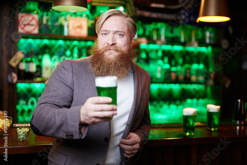 Bearded man with glass of green foaming drink looking at camera in bar on Saint Patrick day