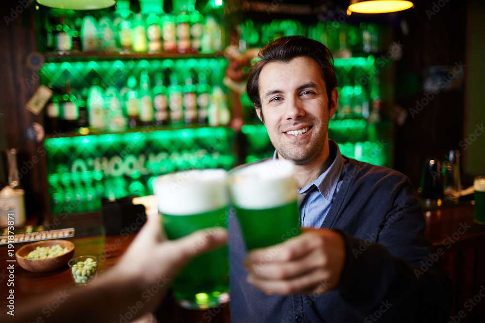 Young smiling man with beer toasting with his friend in pub during celebration of Saint Patrick day