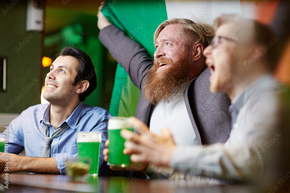 Irish football fans with national flag watching television broadcast in pub by glass of beer
