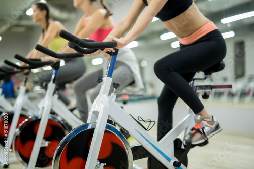 Row of sporty girls sitting on training bikes in sports center and doing exercises for leg muscles