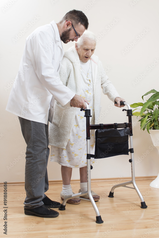 8. Seniorka w białym szlafroku i koszuli nocnej idzie przy pomocy chodzika. Pielęgniarz podtrzymuje i asekuruje starą kobietę.