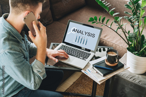 Young man, dressed in shirt, sits at home on couch at coffee table,uses laptop with inscription statistical analysis on screen, talks on his cell phone,calling. Online marketing,education, e-learning.