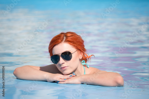 Portrait of a beautiful woman getting out of a swimming pool. beautiful long hair tanned model posing by blue pool water. Outdoor summer portrait of sexy girl in sunglasses. woman in red swimsuit. © Rudkov