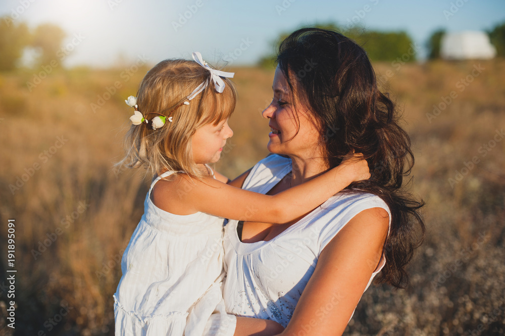 Little blond girl in white dress hugging mom in the rays of a warm summer sunset. Happy smiling child and mother. Little blond girl in white dress hugging mom in the rays of a warm summer sunset. Happy smiling child and mother.