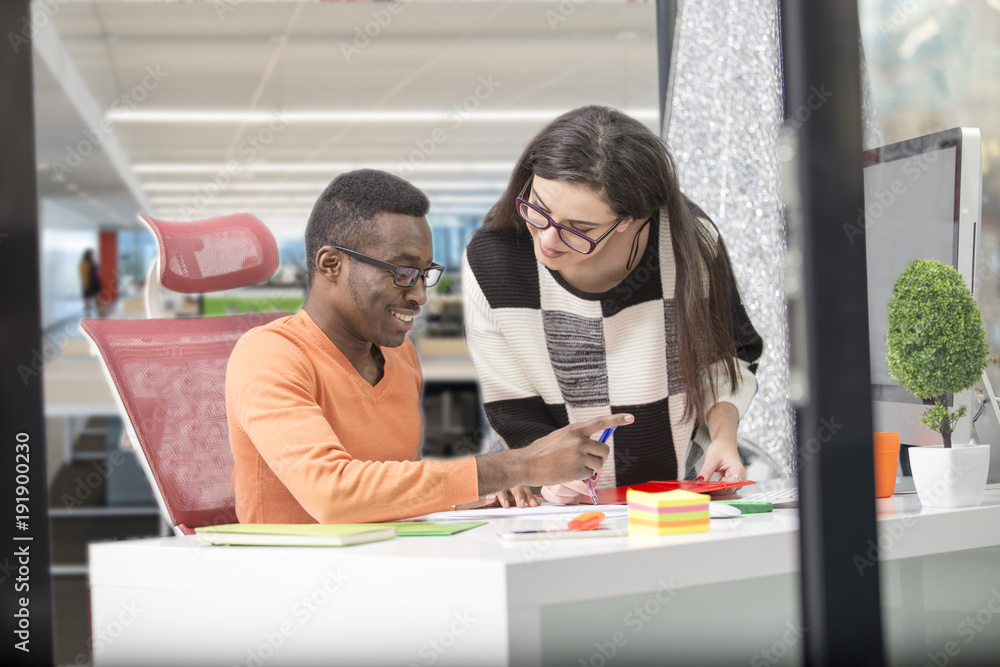 Fototapeta premium Two diverse work colleagues smiling and writing down notes while sitting together at a table in a modern office