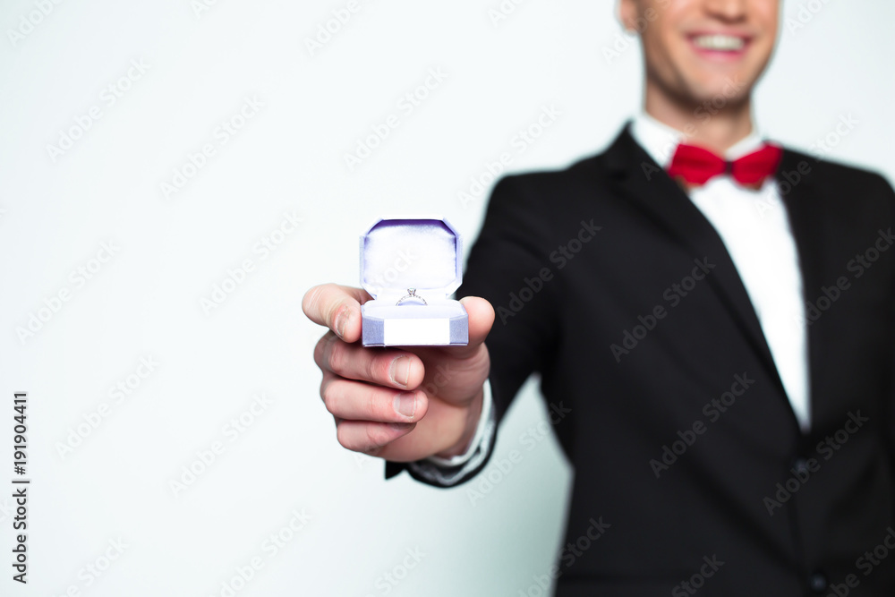 Handsome smiling modern elegant man in a official black suit and a red bow tie is holding a box with an engagement ring and shows in camera. Handsome smiling modern elegant man in a official black suit and a red bow tie is holding a box with an engagement ring and shows in camera.