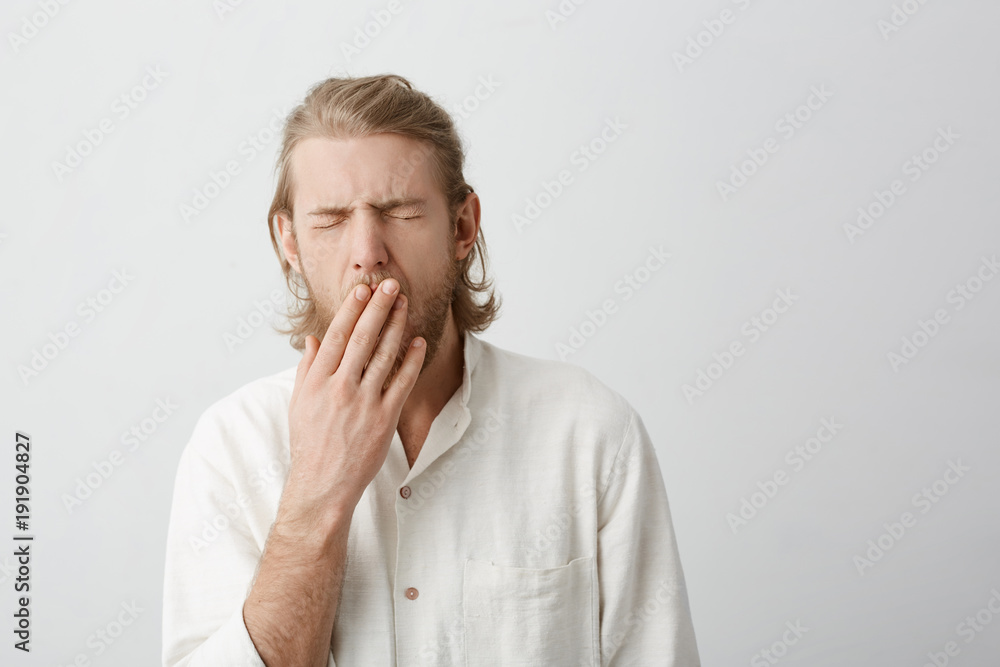 Indoor portrait of young attractive blond male yawning and covering mouth with hands, standing with closed eyes over gray background. Man is tired and exhausted cause he have insomnia Indoor portrait of young attractive blond male yawning and covering mouth with hands, standing with closed eyes over gray background. Man is tired and exhausted cause he have insomnia