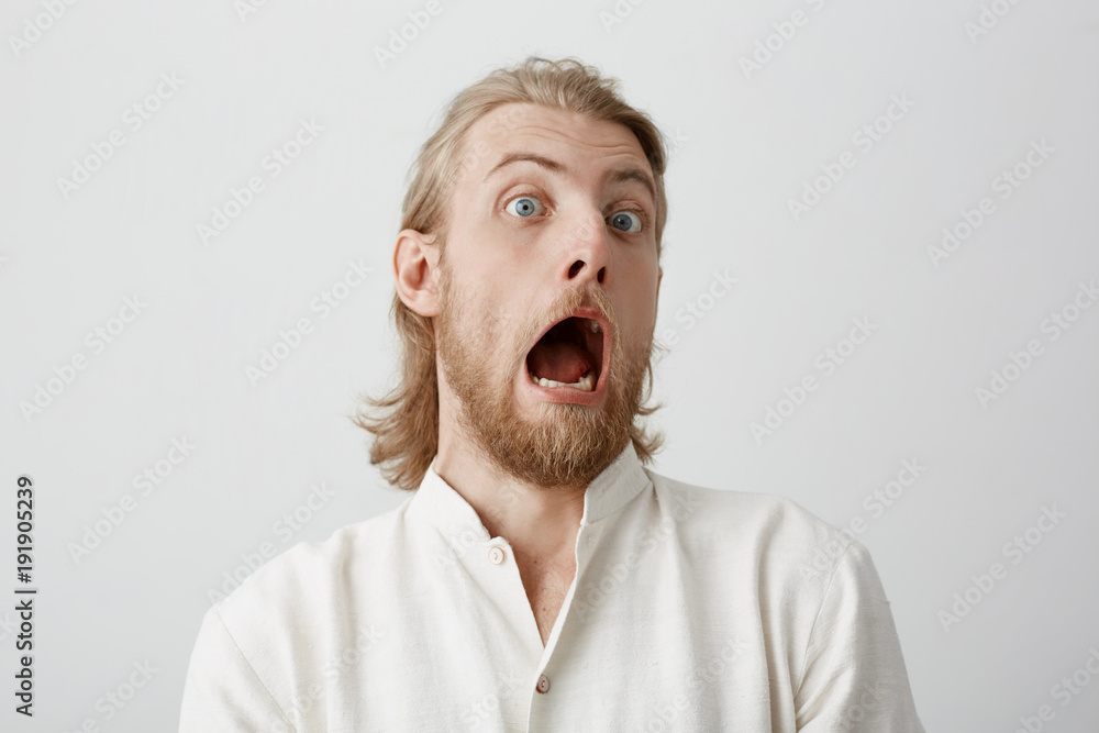 Portrait of funny handsome bearded man with fair hair, expressing shock or being terrified of something, standing with opened mouth and popped eyes over gray background. Guy is pale as if he saw ghost Portrait of funny handsome bearded man with fair hair, expressing shock or being terrified of something, standing with opened mouth and popped eyes over gray background. Guy is pale as if he saw ghost