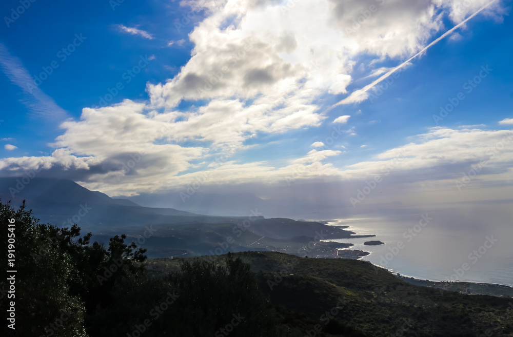 View of the coast and the mountains and a very blue sky with clouds and a vapor trail and the sun highlighting the edge of the ocean