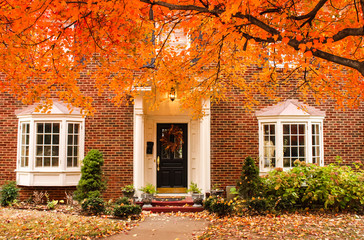 Red brick house entrance with seasonal wreath on door and porch and bay windows on autumn day with leaves on the ground and hydrageas still in bloom - colorful foliage