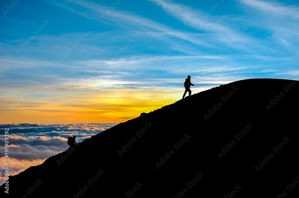 Two people climbing a mountain are almost on the summit. It is sunrise with clouds in the sky. Full of color. 