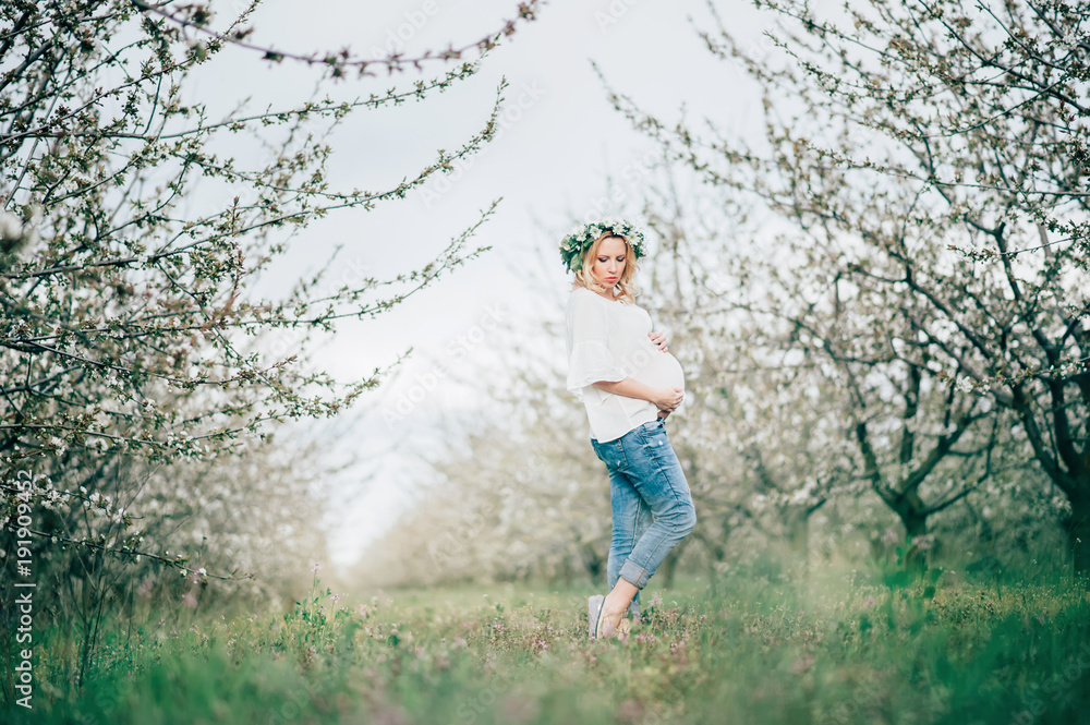 Beautiful young cheerful pregnant woman in wreath of flowers on head touching belly while walking in spring tree garden. Beauty People Motherhood concepts. Happy mom in waiting for baby! Beautiful young cheerful pregnant woman in wreath of flowers on head touching belly while walking in spring tree garden. Beauty People Motherhood concepts. Happy mom in waiting for baby!