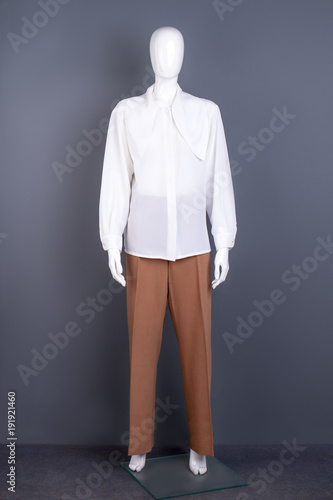 Women white chiffon shirt on mannequin. Full length dummy dressed in long sleeves blouse and brown trousers, grey background.