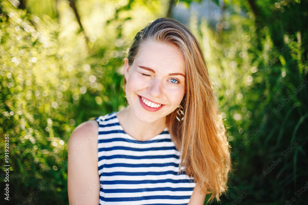beautiful smiling winking white Caucasian girl woman with long blonde hair and blue eyes wearing striped t- shirt outside in summer park among green foliage trees