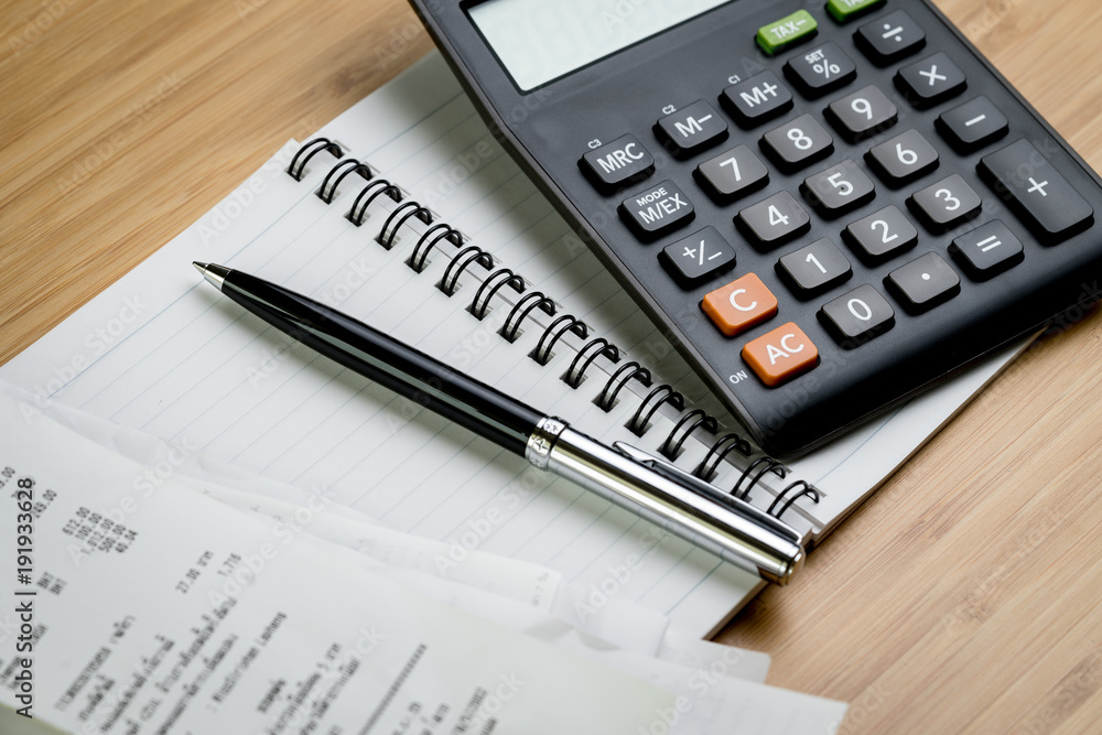 Bill payment or cost calculation concept, black pen on paper notepad with pile of bills and calculator on wood table Bill payment or cost calculation concept, black pen on paper notepad with pile of bills and calculator on wood table