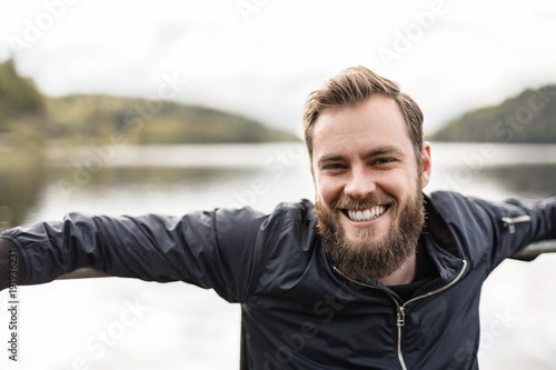 Bearded man sitting down outdoors in front of a lake on a moody day. Wearing a blue jacket looking at camera.