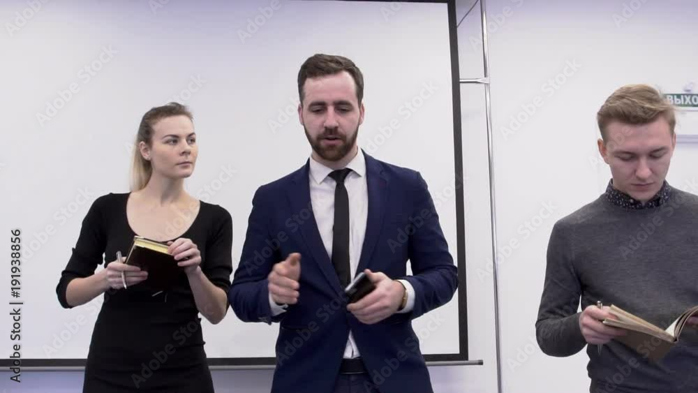 Successful businessman is giving tasks to his iterns in the conference room. Young woman in black dress and blond man in grey jumper are writing the information to their notebooks while going in a row Successful businessman is giving tasks to his iterns in the conference room. Young woman in black dress and blond man in grey jumper are writing the information to their notebooks while going in a row
