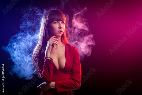 Young dark-haired woman in a red dress posing against a background of red and red smoke from a vape on a black isolated background