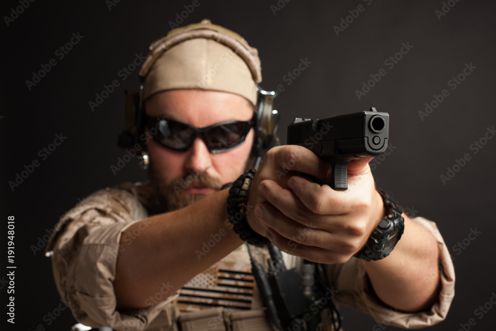 Brutal man in the military desert uniform and body armor standing in a fighting rack and aiming from his gun on a black background. Close-up of the gun barrel directed towards the camera