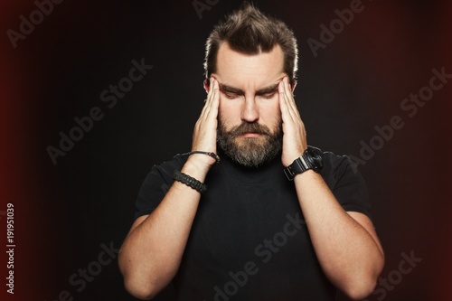 Terrible headache. Frustrated man touching head with hands and making face while standing against black background. It is surrounded by a red glow, symbolizing pain and suffering. Copy space for text photo