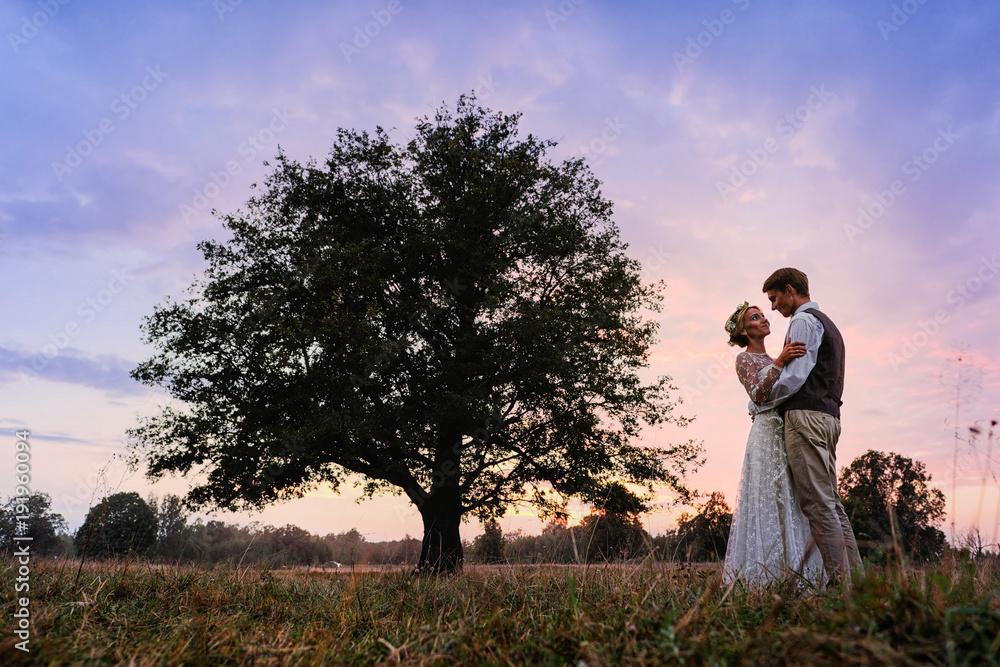 The bride and groom are standing next to a large tree, after the wedding ceremony. Silhouette photo in warm colors.