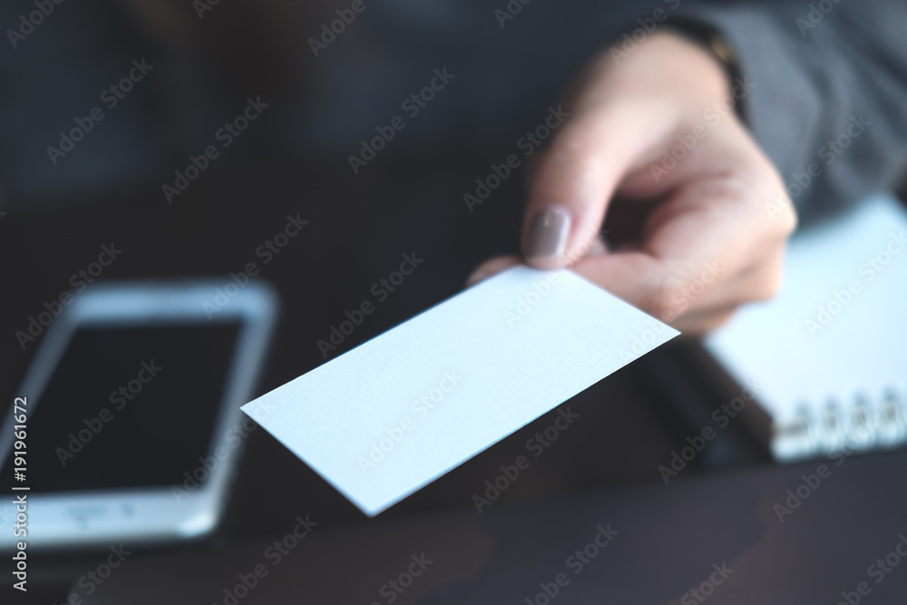 Business woman holding and giving business card to someone with mobile phone and notebook on table in office Business woman holding and giving business card to someone with mobile phone and notebook on table in office