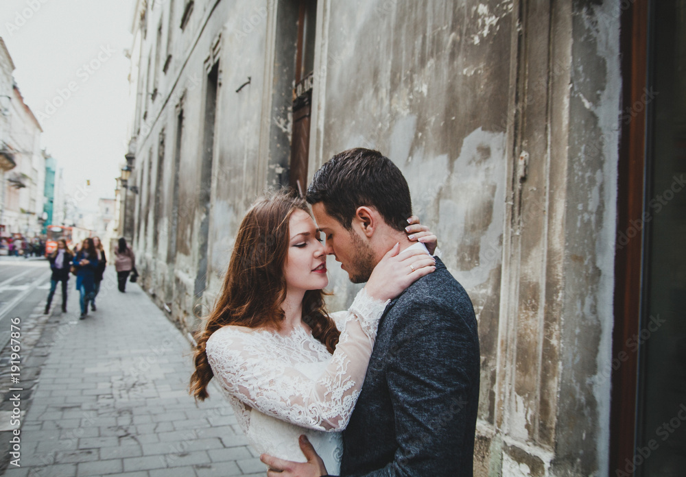 Wedding couple hugs in the old city. Stone walls of ancient town on background. Rustic bride with hair down and groom in grey suit and bow tie. Romantic love in vintage atmosphere street. Love story.