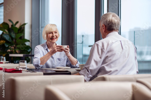 Happy time. Positive pretty mature woman holding cup while smiling and sitting at the table