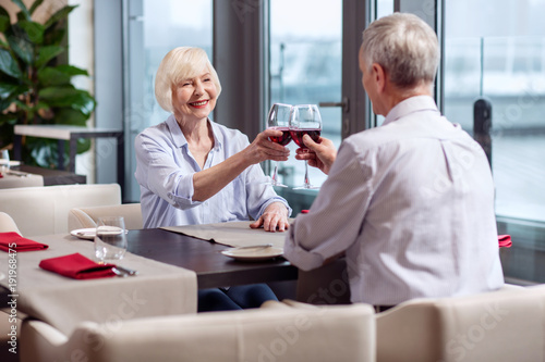 Cheers darling. Happy vigorous mature woman rising glass while smiling and visiting restaurant