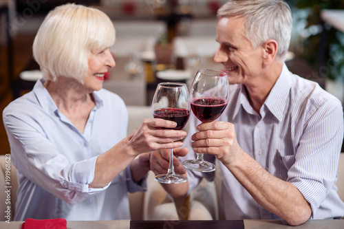 Life together. Energetic jolly mature couple holding glasses of wine while communicating and sitting in profile