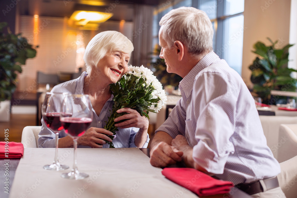 Mature love. Lovely sincere mature woman grinning while holding flowers while man staring at her Mature love. Lovely sincere mature woman grinning while holding flowers while man staring at her