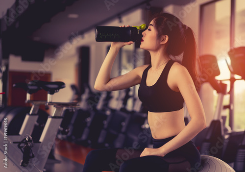 Asian woman drinking protein shake from bottle after workout exercise for muscle at fitness gym healthy lifestyle bodybuilding sitting on fitness ball