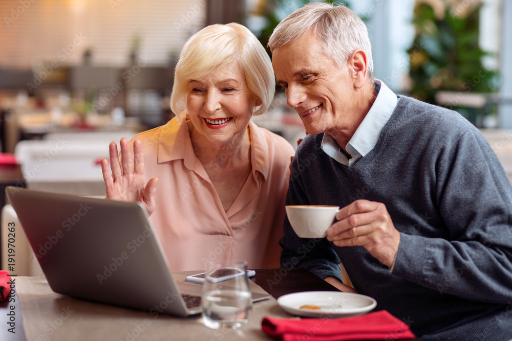 Hi granddaughter. Attractive jolly mature couple sitting at the table and looking at screen while man drinking coffee Hi granddaughter. Attractive jolly mature couple sitting at the table and looking at screen while man drinking coffee