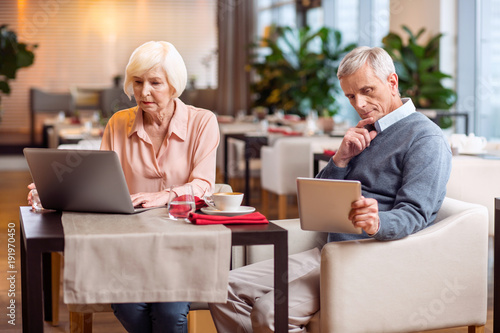 Gadget for everyone. Thoughtful nice mature couple sitting on chairs and looking at screens while using tablet and laptop