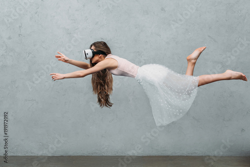 Fototapeta Naklejka Na Ścianę i Meble -  young barefoot woman in virtual reality headset levitating on grey