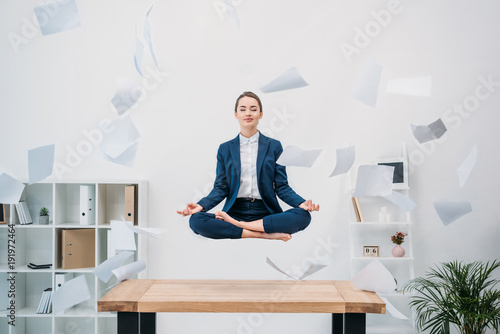 smiling young businesswoman with closed eyes meditating while levitating at workplace with papers