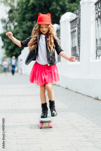 Stylish fashion little girl child riding skateboard on city street. Red hat, black biker jacket.