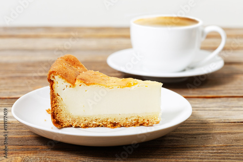 Piece of a cheesecake on a white saucer and cup of espresso on wooden table. Shallow focus.