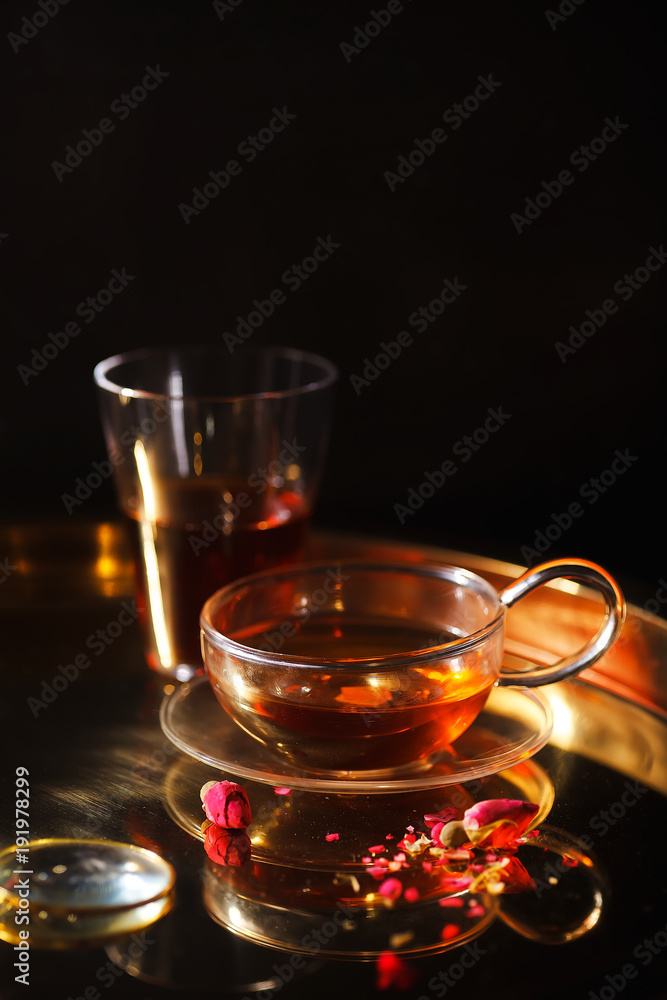 Vertical close-up shot of small glass teapot and cup with hot red tea, dried rose petals, pocket magnifier on golden chain on golden tray. Evening light. Golden color concept. Vertical close-up shot of small glass teapot and cup with hot red tea, dried rose petals, pocket magnifier on golden chain on golden tray. Evening light. Golden color concept.