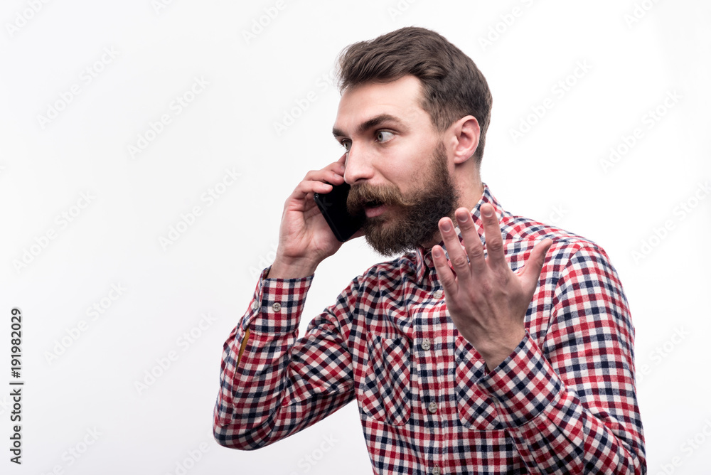 Heated conversation. Handsome bearded man in a checked shirt talking on the phone emotionally and gesticulating, seeming disagreeing with his interlocutor Heated conversation. Handsome bearded man in a checked shirt talking on the phone emotionally and gesticulating, seeming disagreeing with his interlocutor