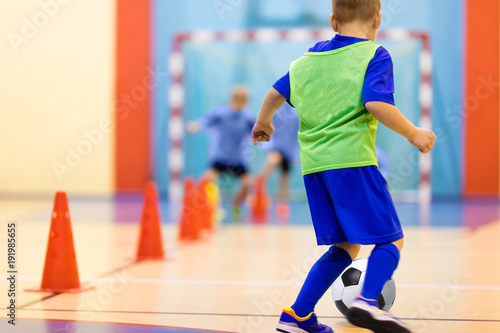 Soccer training dribbling cone drill. Football futsal training for children. Indoor soccer young player with a soccer ball in a sports hall. Player in blue uniform. Sport background.