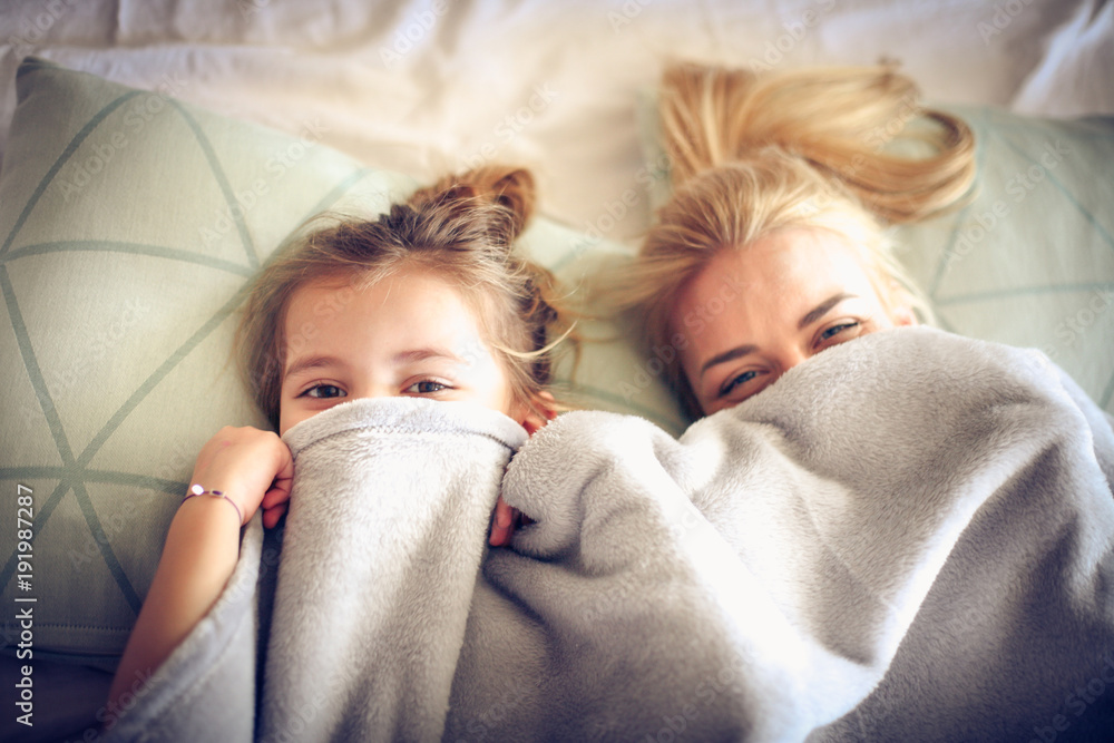 Portrait mother and daughter hide under blanket on a bed. - obrazy, fototapety, plakaty 