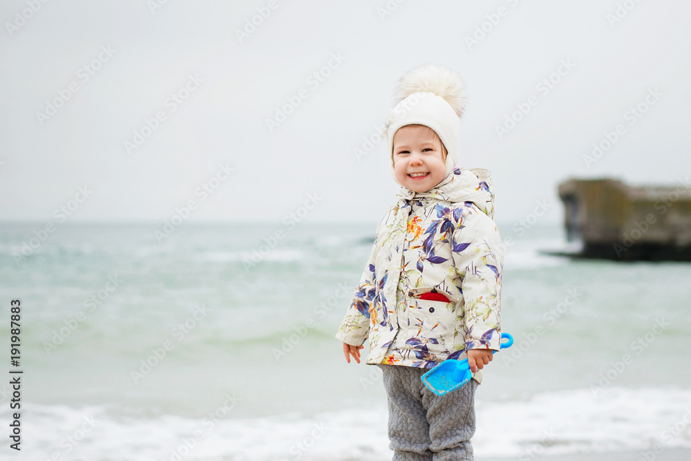 Cute little girl playing on the sandy beach. Happy child wearing warm floral print jacket, pom pom hat and scarf playing outdoors on fall, winter or spring day. Cute little girl playing on the sandy beach. Happy child wearing warm floral print jacket, pom pom hat and scarf playing outdoors on fall, winter or spring day.