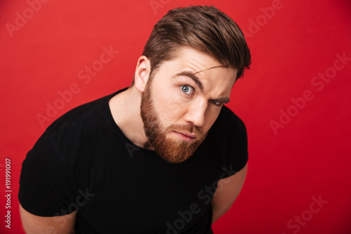 Portrait of muscular man 30s with suspicious look wearing black t-shirt posing on camera with raising eyebrow, isolated over red background
