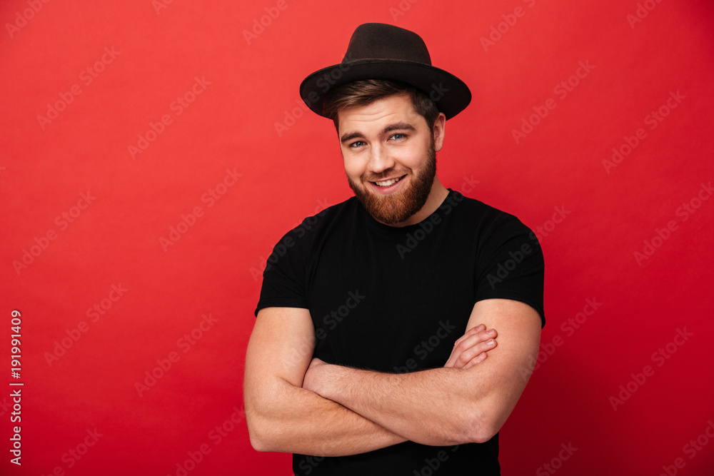 Portrait of irresistible stylish man 30s wearing black t-shirt and hat smiling on camera and posing with hands crossed, isolated over red background