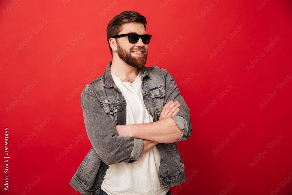Wizerunek mody młody człowiek jest ubranym okulary przeciwsłonecznych i drelichowy ono uśmiecha się i pozuje z rękami składać, odizolowywający nad czerwonym tłem <span>plik: #191991455 | autor: Drobot Dean</span>
